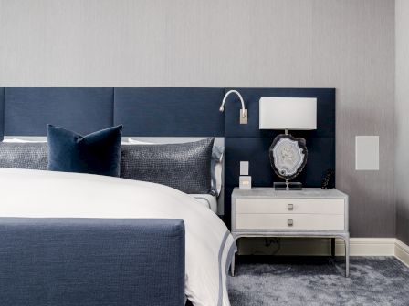 The image shows a modern bedroom with a blue upholstered headboard, a white bed, a nightstand with a lamp, and a decorative item.