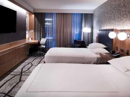 This image shows a modern hotel room with two double beds, a wall-mounted TV, a desk with a chair, and contemporary lighting and decor.