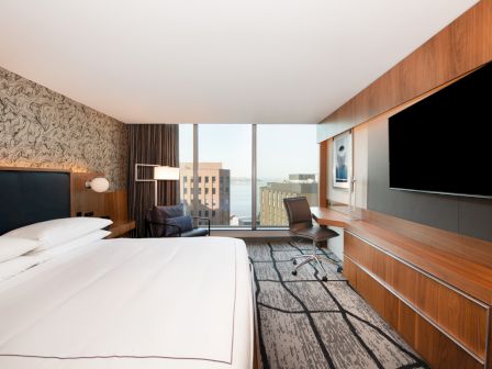 This image depicts a modern hotel room with a large bed, a desk with a chair, a flat-screen TV, and a window offering a city view.