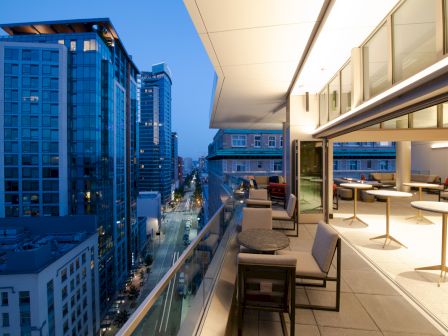A modern balcony view overlooking a cityscape, featuring outdoor seating and tables, with high-rise buildings in the background.