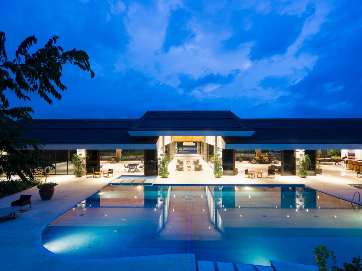 A modern building and illuminated swimming pool at night, with lounge chairs and open seating areas visible under a cloudy sky.