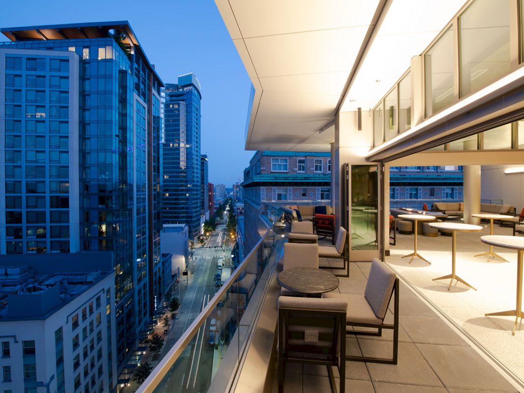 An outdoor balcony area overlooks a city street lined with modern buildings at dusk, featuring seating and tables on the patio.