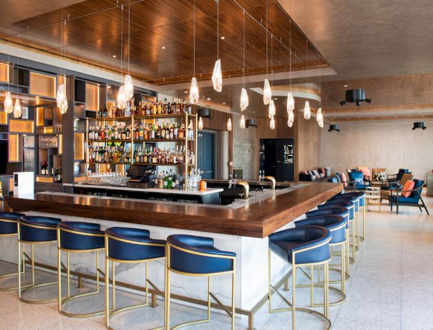 This image shows a modern bar with a wooden counter, blue cushioned bar stools, hanging lights, and a fully stocked liquor shelf in a spacious setting.