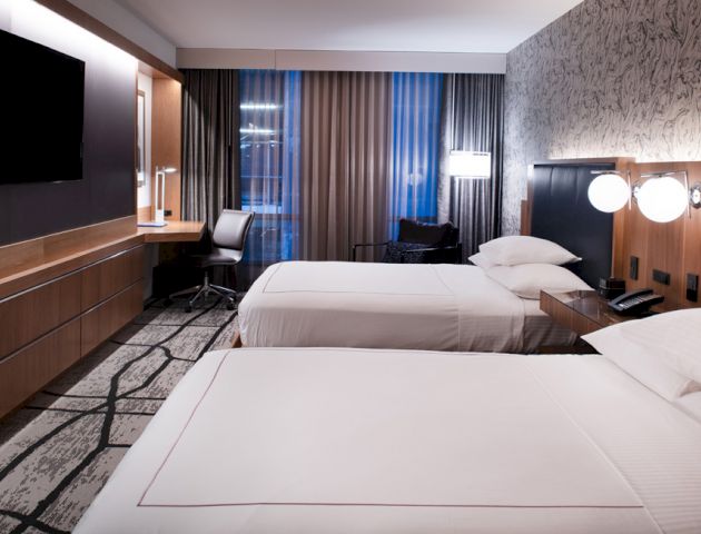 This image shows a modern hotel room with two beds, a wall-mounted TV, a desk with a chair, and stylish lighting and decor.