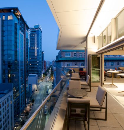 A modern cityscape at dusk featuring a high-rise balcony with outdoor seating, overlooking a street lined with tall buildings.