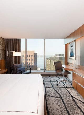 A modern hotel room with a large bed, wall-mounted TV, desk, chair, and large window offering a cityscape and water view, ending the sentence.