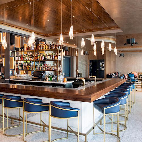 A modern bar with high blue stools, a well-stocked liquor shelf, and pendant lights, adjacent to a lounge area with seating.