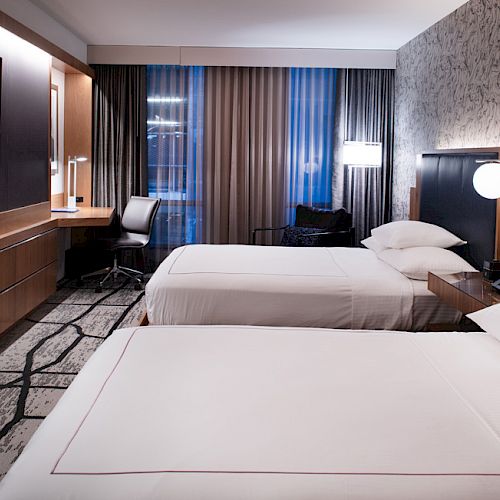 The image shows a modern hotel room with two neatly made beds, a flat-screen TV, a work desk and chair, and contemporary decor.