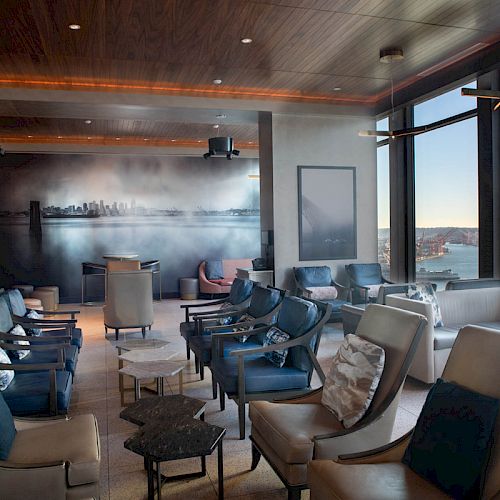 A modern lounge with armchairs, tables, a large window with a waterfront view, and a wall mural of a city skyline in the background.