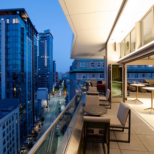 The image shows a modern balcony with seating areas overlooking a city street and high-rise buildings in the evening.