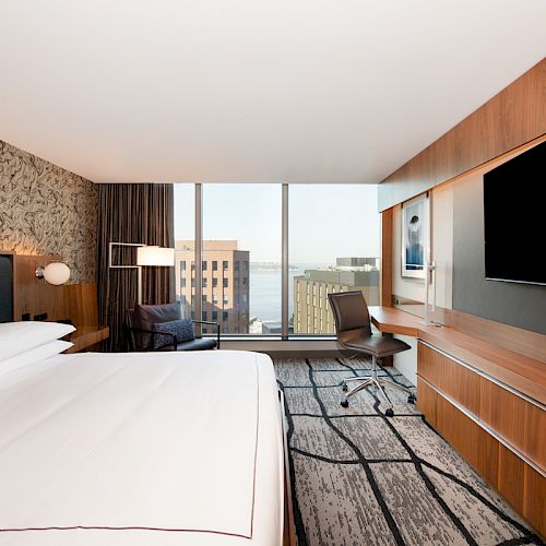 A modern hotel room with a large bed, desk, chair, wall-mounted TV, and a window offering a city view is shown in this image.