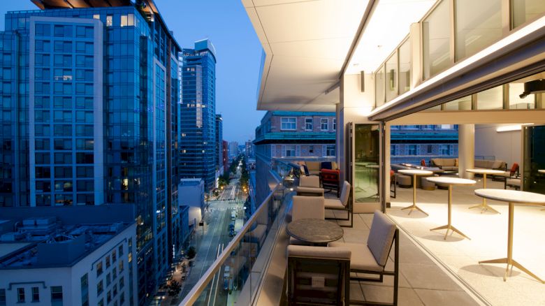 A modern city skyline at dusk with a view from a building's balcony, furnished with chairs and tables, overlooking a street with lights.