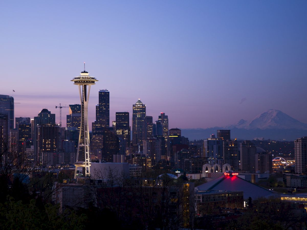 The image shows the Seattle skyline at dusk, featuring the iconic Space Needle and Mount Rainier in the background, with illuminated city buildings.