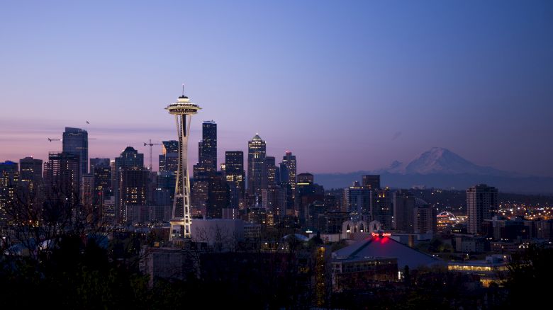 The image shows the Seattle skyline at dusk, featuring the iconic Space Needle and Mount Rainier in the background, with illuminated city buildings.