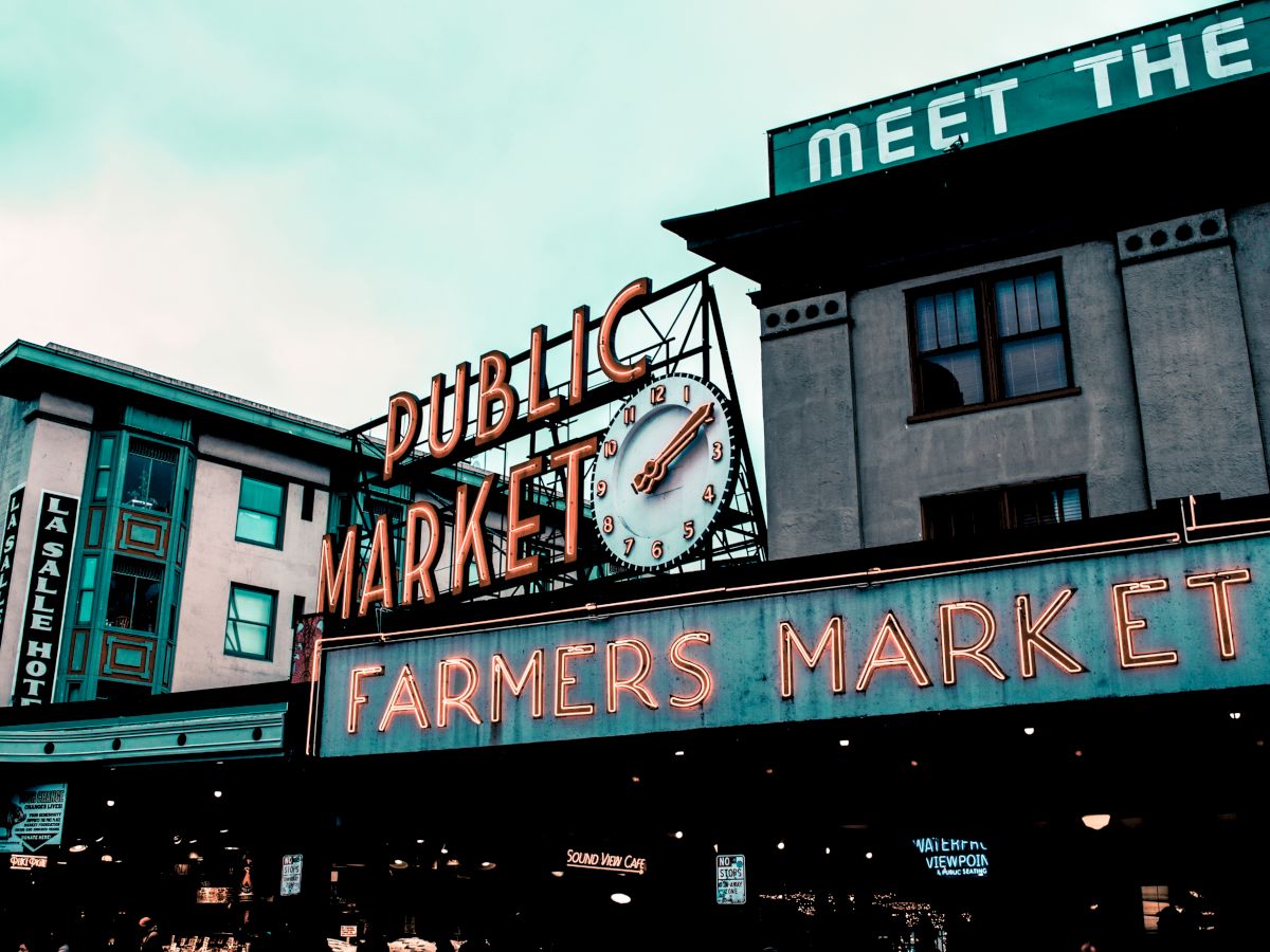 This image shows the entrance sign of a public market and farmers market, with the word "HOTEL" visible on a building in the background.