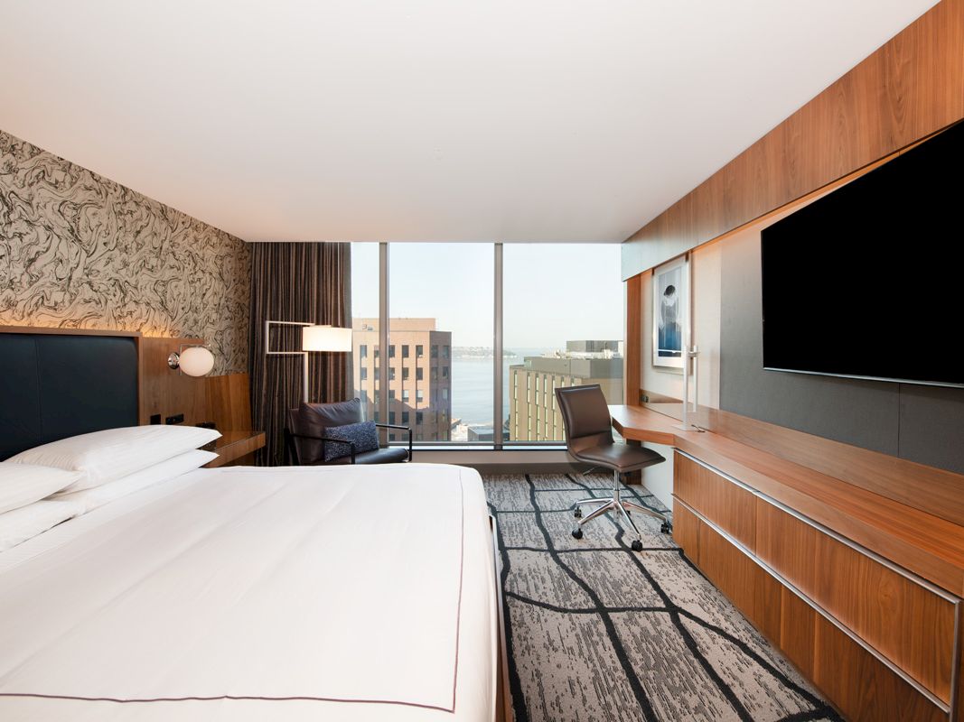 A modern hotel room features a large bed, a desk with a chair, a flat-screen TV, and windows offering a city and water view.