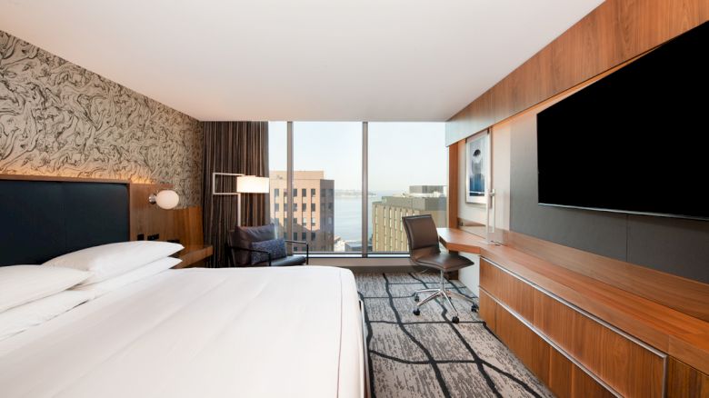 A modern hotel room features a large bed, a desk with a chair, a flat-screen TV, and windows offering a city and water view.
