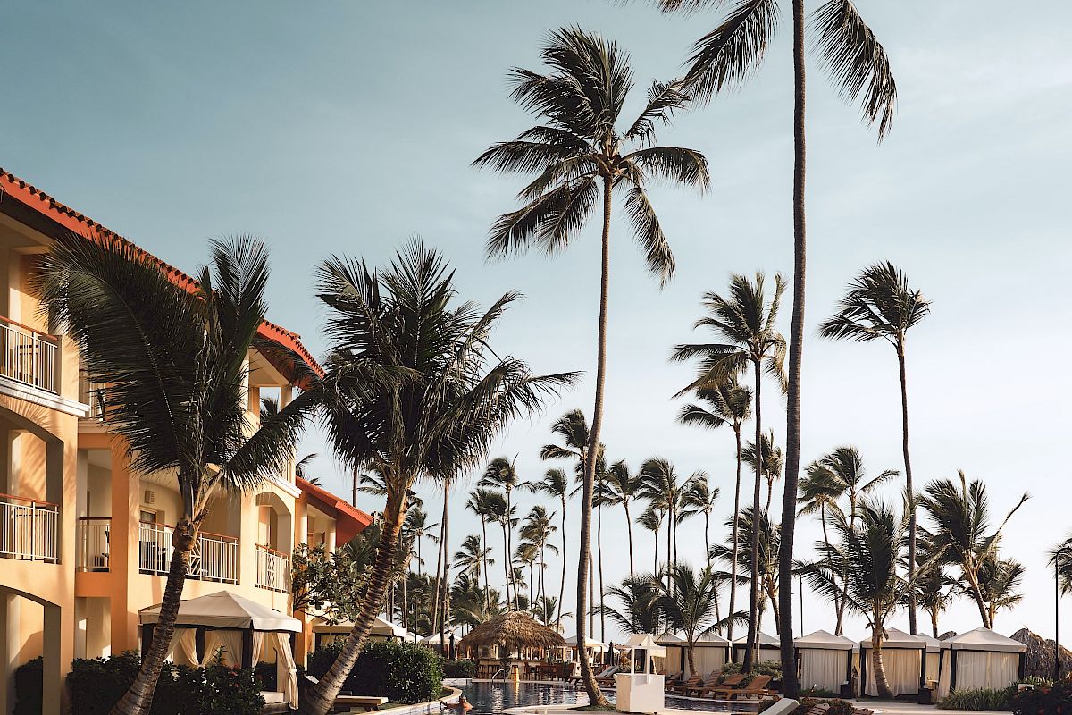 A luxurious poolside scene with wooden lounge chairs, tall palm trees, and a building with balconies under a clear sky.