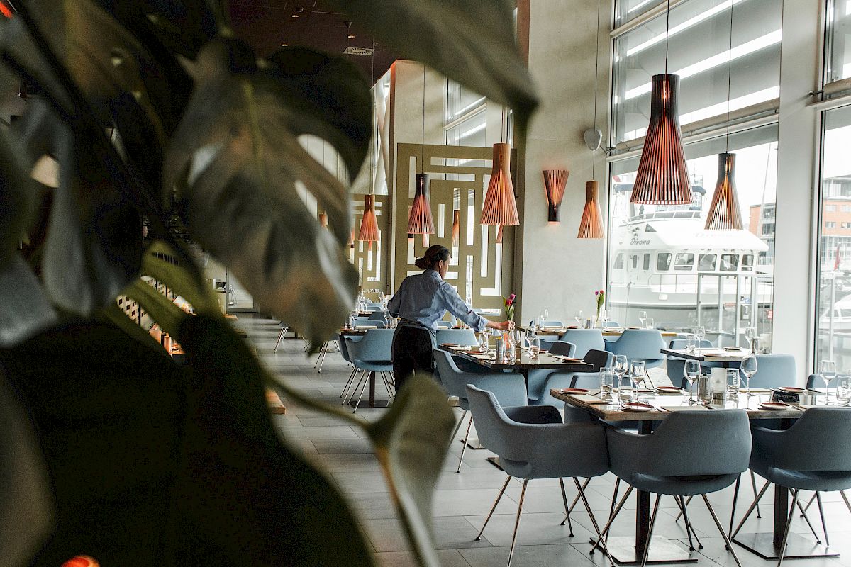 A person is arranging tables in a modern restaurant with stylish lighting and large windows, viewed through some indoor plants.