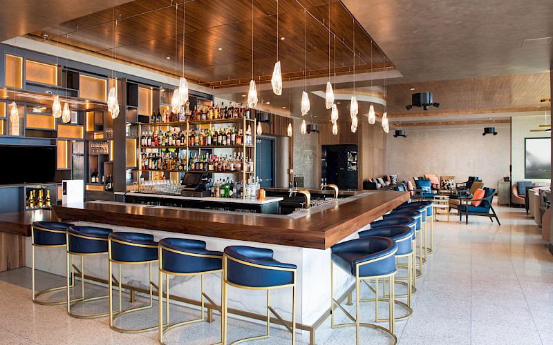 This image shows a modern bar with blue and gold barstools, a well-stocked bar, hanging lights, and seating areas in a stylish and spacious environment.