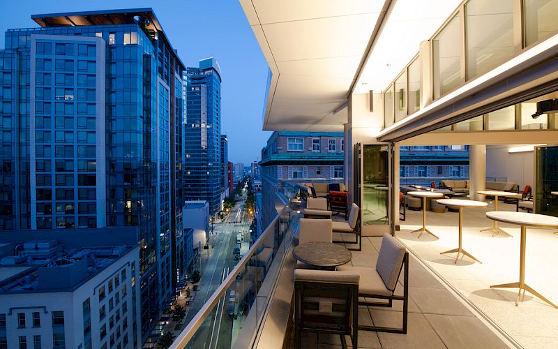 A modern rooftop balcony with seating and tables overlooks a cityscape filled with tall buildings in the evening.