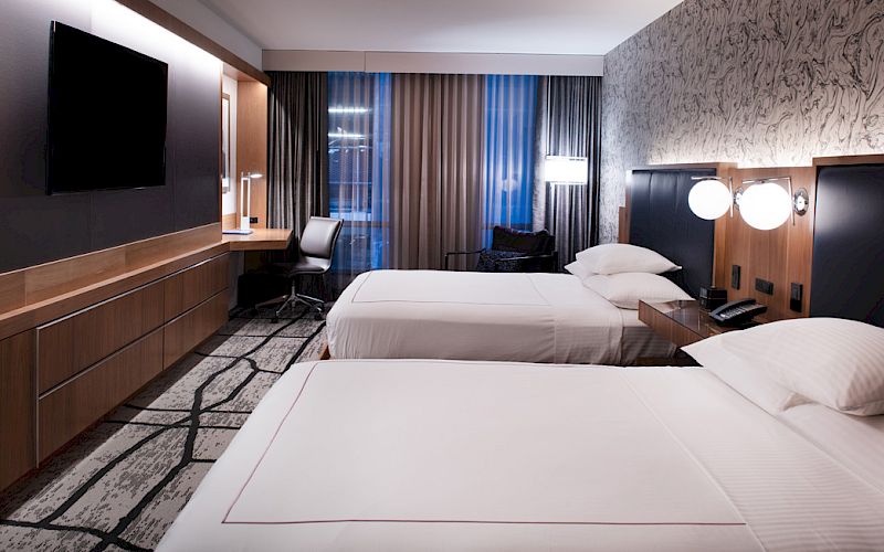 A modern hotel room with two neatly made beds, a wall-mounted TV, a work desk, and stylish decor, illuminated by soft lighting and natural light.
