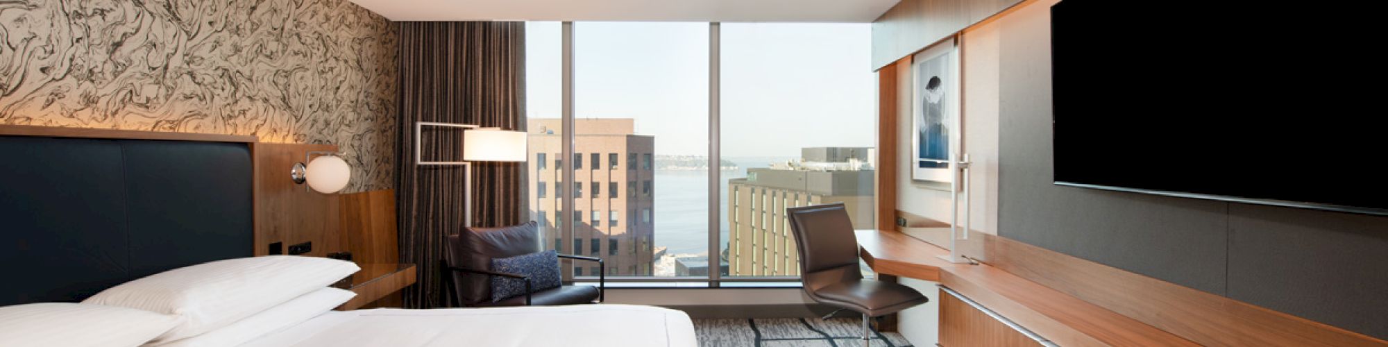A modern hotel room with a large bed, wall-mounted TV, desk, and a window offering a cityscape view, and stylish design elements throughout.