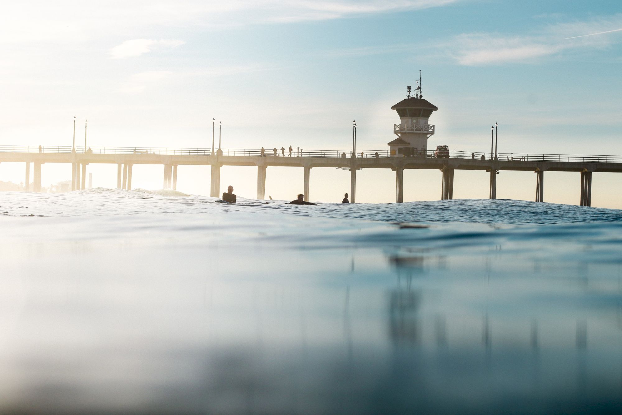 The image shows a pier extending over a calm ocean with silhouettes of people on the pier and a lifeguard station visible against the sky.